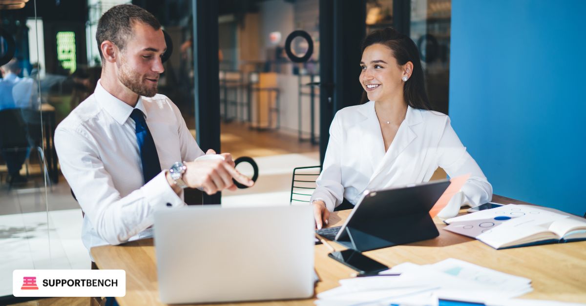 A business professional (wearing business attire) discussing and comparing two helpdesk platforms on a laptop with a colleague in a modern office setting.